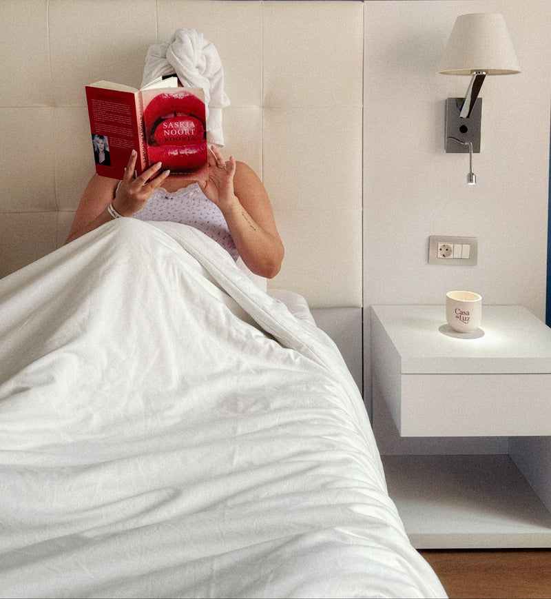 Person lying in bed reading a red book with a white nightstand and lamp in the background, CANDLE LIT