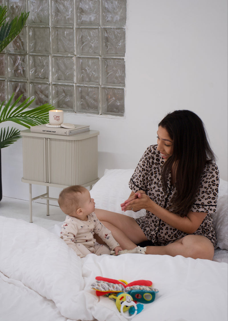 Woman and baby sitting on a bed with toys around, in a room with a glass block wall.